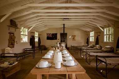 Colour photograph showing interior of wooden hut with trestle tables in centre and beds around sides