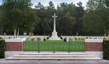 Colour photograph showing a cenotaph behind a metal gate with rows of well tended gravestones in the background