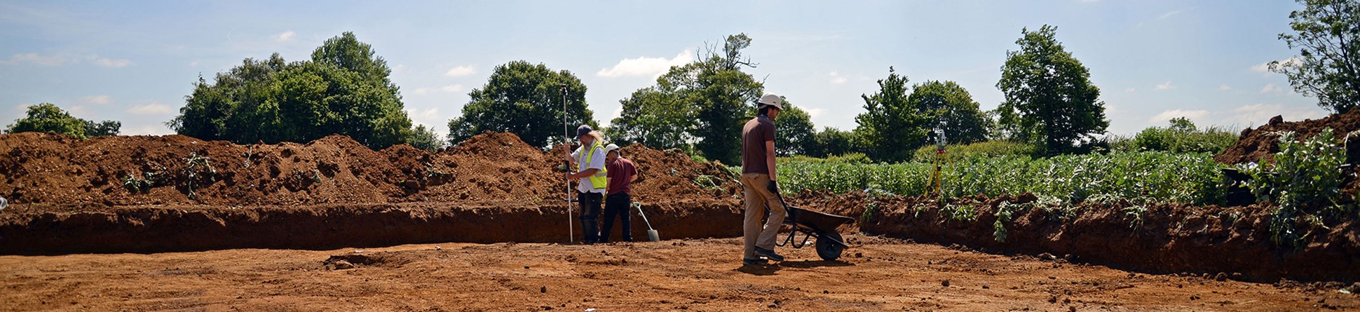 Photo of wide view of excavation trench with spoilheap at rear. In the trench is a man with a wheelbarrow and two surveying