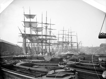 In the A black and white Victorian photo of London docks. Small boats are crowded together in the foreground. There are old, masted sailing ships in the background.