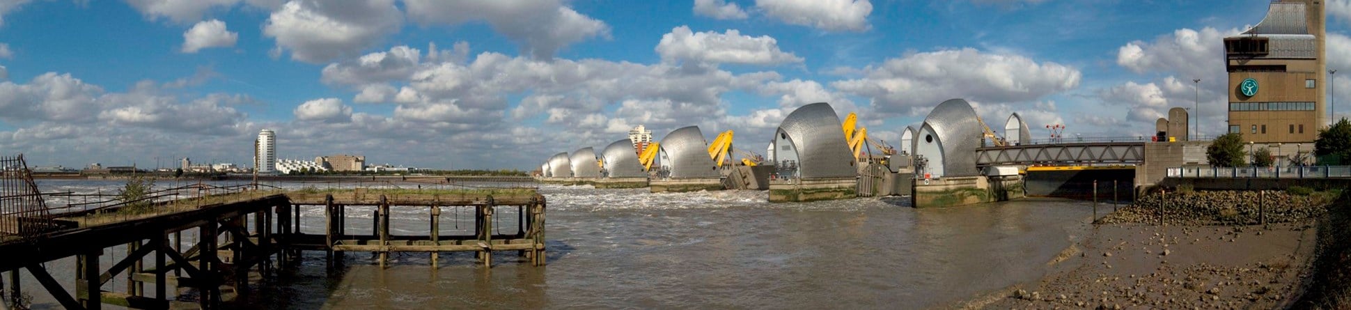 A wide angled photograph of a jetty and rocky foreshore in front of the Thames Barrier, London.
