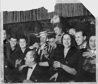 The photograph shows Ian Board, Colony Room bartender and later proprietor (2nd), David Archer, bookseller (4th, seated), Martita Hunt, actress (7th), Muriel Belcher (8th), Carmel Stuart, Muriel Belcher's lover (11th)