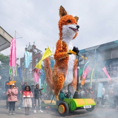 A large fox puppet in a smokey street scene, surrounded by children waving colourful banners.