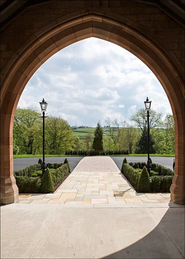 A photograph of a view looking out at a path, road, ornate lampposts, countryside and a cloudy sky through a stone archway