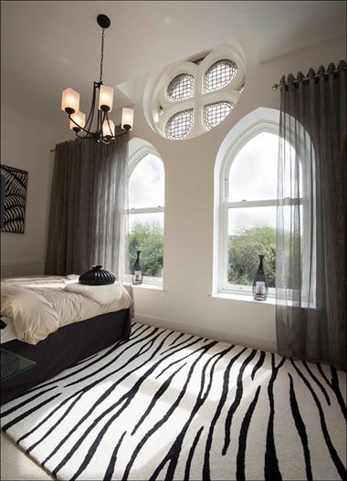 A photograph of a bedroom with zebra-style carpet, a bed, curtains and arched windows
