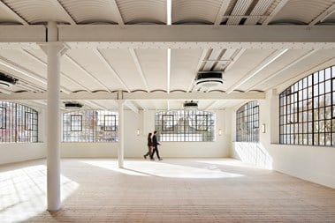 An interior photograph of an empty open-place office space with white girders and large, latticed windows. Two people are walking around the room in the background.