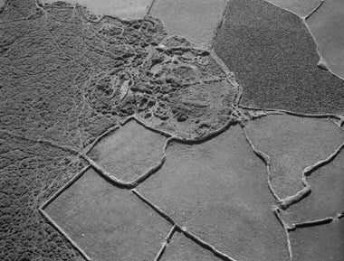 Black and white oblique photograph that reveals a patchwork quilt-like view of irregular-shaped enclosures bounded by low stone walls. Several of the enclosures appear relatively bare of vegetation, whilst one shows signs of a growing crop. To the left side of the photograph is an area of heath lined with narrow, winding tracks. Just above the centre of the photograph is an overgrown archaeological feature enclosed by a roughly circular wall. Inside the enclosure there is evidence of the remains of straight and curved walls.