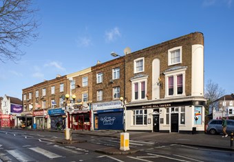 Three-storey brick row of shops and a pub viewed across a road.
