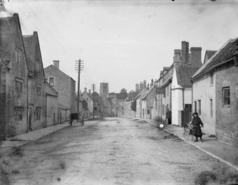 A street view looking towards the church tower, Northleach, Gloucestershire. In the foreground a young girl watches the cameraman at work. She is pulling a child in what is believed to be an invalid carriage. 1901. 