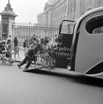 A man in a wheelchair is pushed up a ramp into the back of a waiting van run by The Not Forgotten Association, a charity for disabled army veterans, in front of the railings of Buckingham Palace, 1950s.