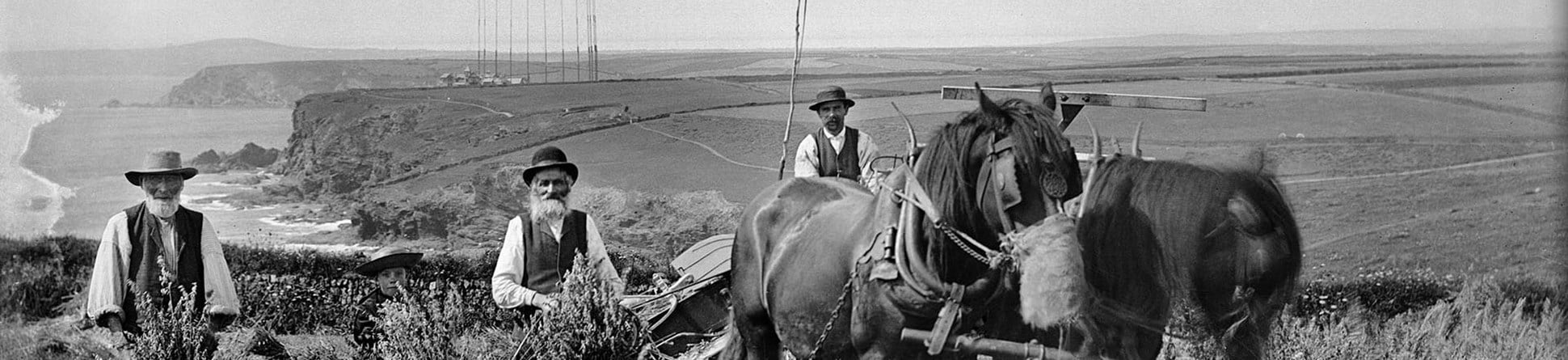 Black and white archive photograph of three farm workers and a horse drawn vehicle posing for the camera in a field, with radio masts, cliffs and coasts in the background.