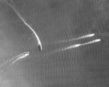 Detail from a black and white vertical aerial photograph, showing warships at sea. A vessel towards the centre of the image sails towards the bottom, its wake trails from the top-left corner of the image. Three vessesl sail towards the right edge of the image. Another vessel sails from the left towards the first vessel. 