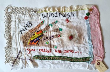 Fabric with white and coloured hands entwined with the words 'Windrush, NHS angel, you called we came' embroidered on it.