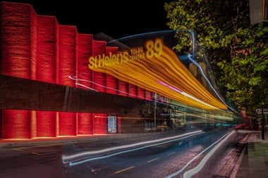 A photograph at night of a building lit up red and a stylistic light trail from a retreating bus reading 'St Helens 89'
