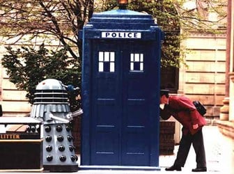 A blue police box on a street, with a dalek on its left and a man in a red coat peering into the box on its right