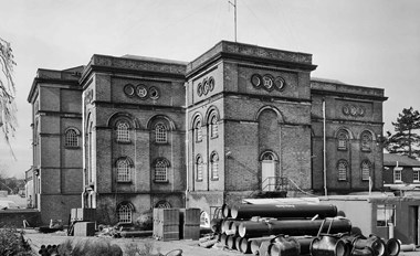 Black and white photo of four-storey pumping station with large piping stacked up outside