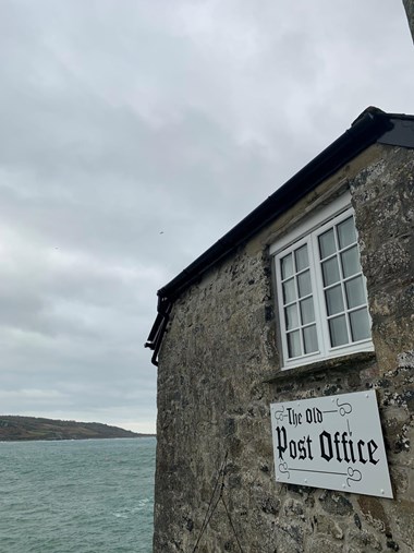 A photograph of an old stone building with the sea in the background. A sign on the building reads ’The Old Post Office’.