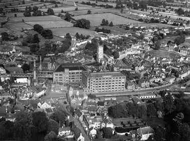 Black and white oblique aerial photograph of a town centre. The centre of the photograph is dominated by multi-storey factory buildings, their adjacent tall chimneys, and the square tower of the parish church. Streets of houses and commercial buildings fill much of the photograph. Rectilinear fields bounded by hedges and trees extend into the distance.