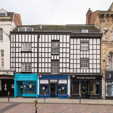 Looking across a high street to a three-story timber-framed building. Its ground floor has a row of shopfronts. Its upper floors and roof show the need for repair. 