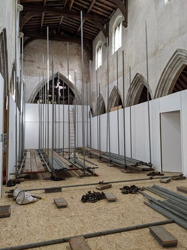 White hoardings surround upright scaffolding poles on a chipboard floor in a church.