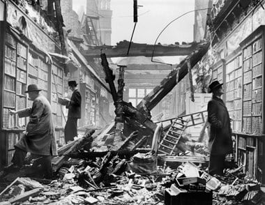 Archive photograph showing three men calmly browsing books on surviving library shelves amid the rubble of a roofless shell