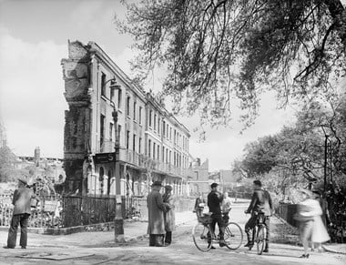 Archive black and white photograph showing people in the road with ruins showing behind a standing street facade.