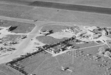 A black and white oblique aerial photograph showing aircraft parked at a dispersal area on the edge of an airfield. Part of a perimeter road and runway can be seen across the top of the image. Adjacent to one hardstand are farm buildings.