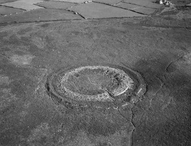 Black and white oblique aerial photograph taken at low altitude showing a circular multivallate hillfort set in a stretch of heath. Two concentric banks are discernible. The outer one is covered in vegetation, with relatively little stone showing. The inner one shows considerably more exposed stone, including surviving walling. There is a narrow path running through both banks and through the centre of the inner enclosure. A pair of upright stones mark the entrance to the inner enclosure. At the top of the photograph a hedge separates the heath from enclosed fields of pasture. 