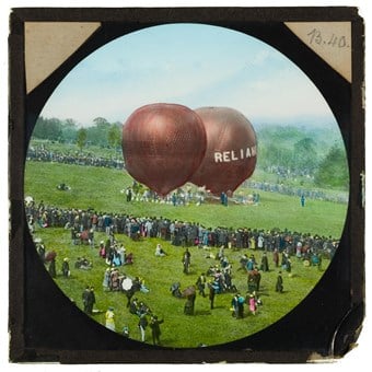 A hand-coloured slide showing Shadbolt's balloon 'Reliance', and a second balloon preparing to take off from the grounds of Alexandra Palace, with large crowds watching the event