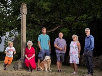 A colourful group of six people and one dog pose next to the Village Cross; an approximately-three meter high stone monument. 