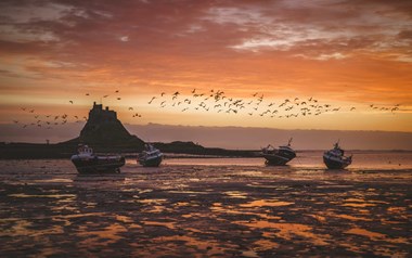 A photograph of four fishing boats anchored on a beach with a flock of birds flying above and the silhouette of a castle set upon a hill in the background.
