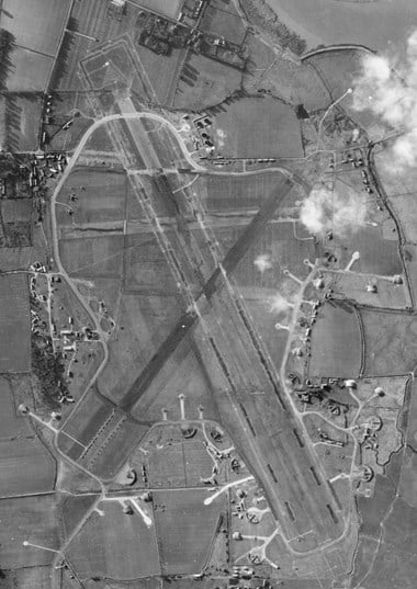 A black and white vertical aerial photograph showing an airfield with three runways. Dispersal areas with hardstands protrude from an irregular perimeter road.