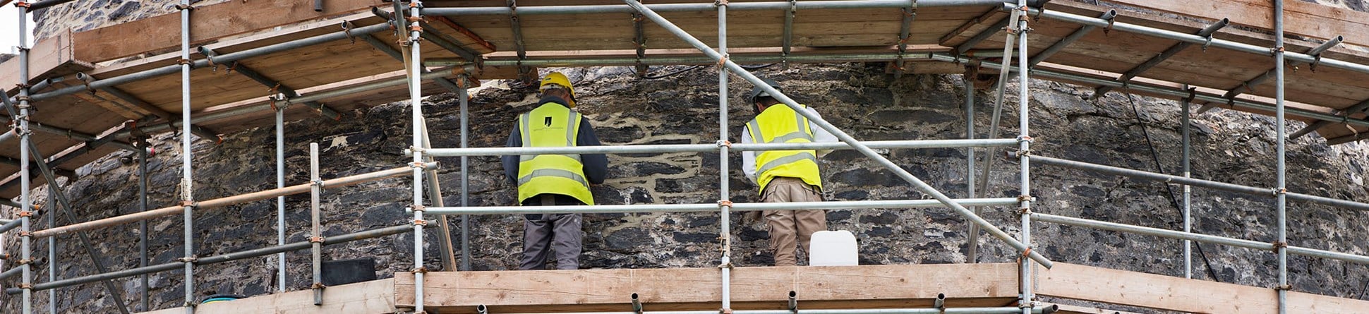 A photograph from behind of two individuals within scaffolding working on a stone wall.