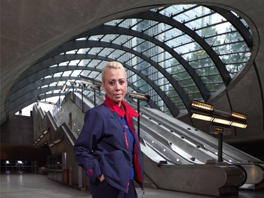 Woman in blue and red London Underground uniform standing at the bottom of the escalators at Canary Wharf Tube station.