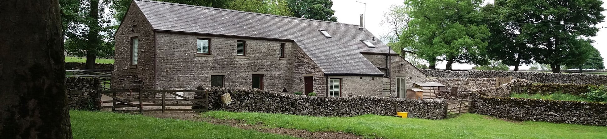 Stone farm building with grey slate tile roof set in fields bordered by dry stone walls with large trees dotted around and a dry stone wall running in front of the house.
