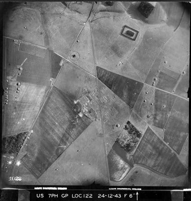 A black and white vertical aerial photograph of a rural landscape featuring a patchwork of irregular fields, tracks, roads and several circular prehistoric earthworks.