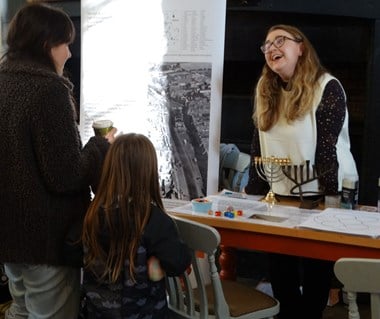 A woman and a young girl interacting with a presenter at a stand with learning materials about Jewish heritage including two menorahs.