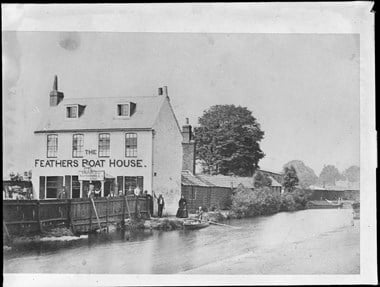 View across a canal to a boat house. A rowing boat with 2 passengers is tied up at the bank. A woman stands next to the boat on the bank and other people are standing outside the boathouse.