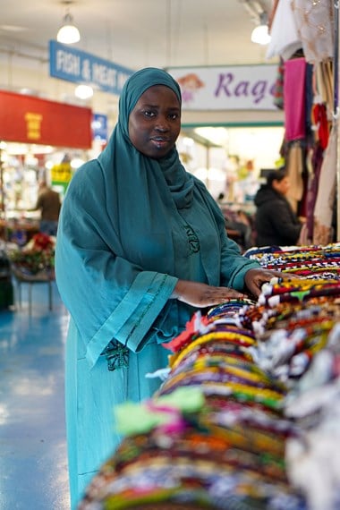 A woman stands with hands on a display of fabrics, looking into the camera. Behind her there are signs and customers of other businesses in the arcade.