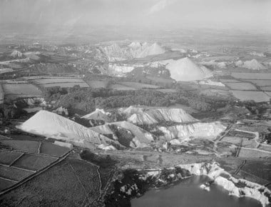 Black and white oblique aerial photograph of china clay works in a landscape of fields bordered with stone walls and hedges, and patches of woodland. The right foreground includes part of a water-filled pit. Stretching across much of the middle and background of the photograph is evidence of excavation and mounds of waste, making the landscape appear filled with miniature white mountains.