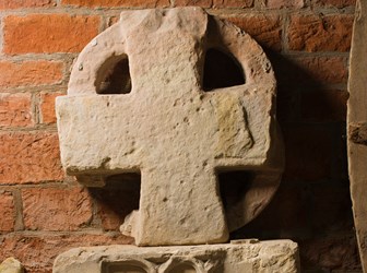A large sandstone cross rests against a red brick wall, on top of a block carved with birds.