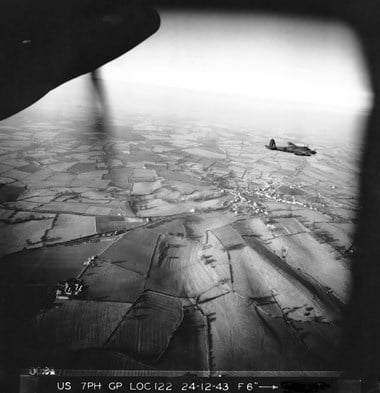A black and white oblique aerial photograph showing a military aircraft flying over a rural landscape. A settlement is visible amongst a patchwork of fields. In the top-left of the image, the spinner and propeller of the aircraft carrying the camera is visible.