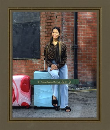 A colour, framed photo of a young woman sitting on a large, blue suitcase. Adjacent is a smaller suitcase. The backdrop is the corner of a brick building. Across part of the lower portion of the photograph is a label that reads: 'Salehasultana Syeo'. 