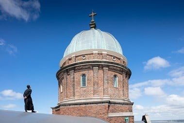 A priest in cassock stands on the roof of a church, looking up at a large brick-built, copper dome.
