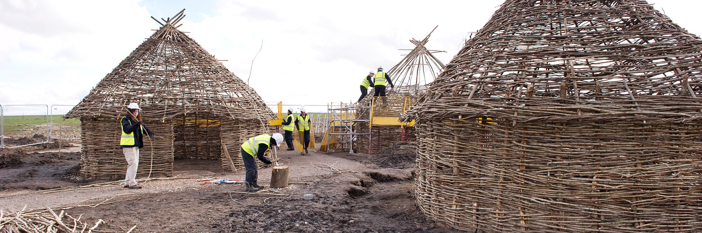 Ground-level image of 6 people in high-visibility clothing and protective helmets building Neolithic-style huts.