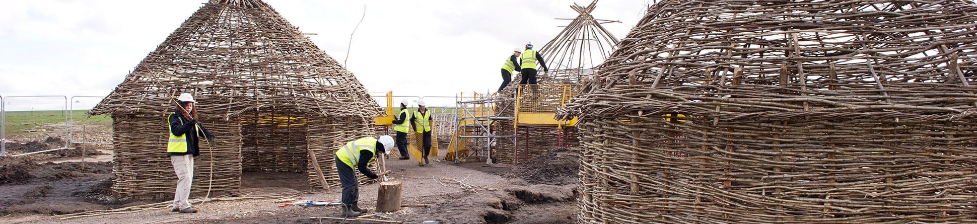 Ground-level image of 6 people in high-visibility clothing and protective helmets building Neolithic-style huts.