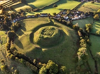 A photograph taken from the air of the earthworks of a large motte and bailey castle, with trees, a road and cottages running along the perimeter.