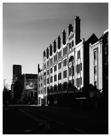 A black and white photograph of a four-storey building with an empty road in the foreground.