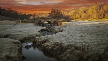 A photograph of a stone bridge set in a frosted rural landscape, with a flowing stream in the foreground.