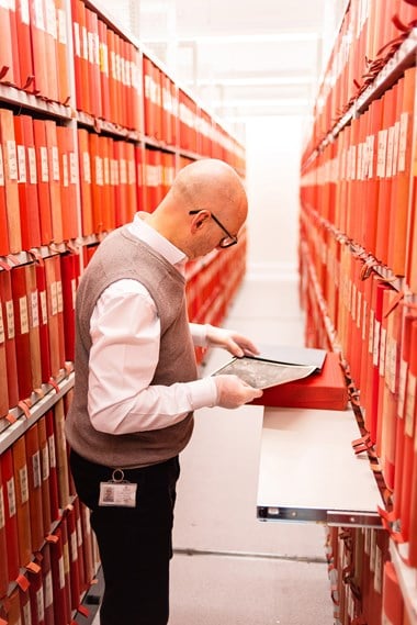 A colour photo of a man at work in an archive storage area. The man stands at a pull-out shelf between roller-racking containing red box files. His gloved hands are carefully returning a photographic print to its sleeve.